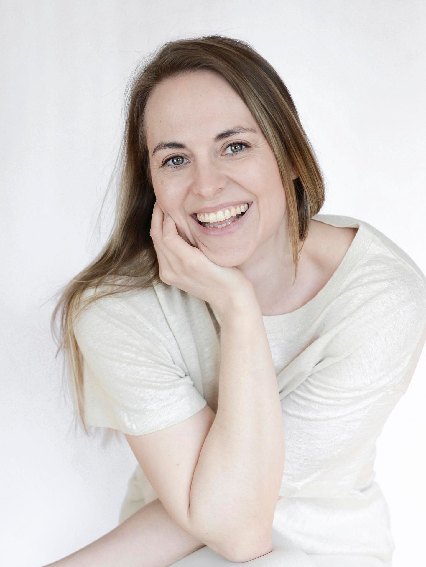 A woman with long brown hair smiles brightly, resting her chin on her hand. She wears a light-colored shirt. The background is plain white. The image shows only her upper body in a straightforward portrait shot, with no products or packaging visible. There are no labels, typography, or additional props present in the scene.
