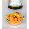 Swanson Curcumin Complex; pieni lasikulho täynnä suuria, oransseja kurkumakapseleita seisoo valkoisella pinnalla; kapselit soikeita ja kiiltäviä; taustalla muovipullo vihreä-valkoisella etiketillä lihavoitua groteskitekstiä, sanat eivät täysin luettavissa epätarkkuuden vuoksi; kuva hyvin valaistu, ei ihmisiä tai käsiä näkyvissä.