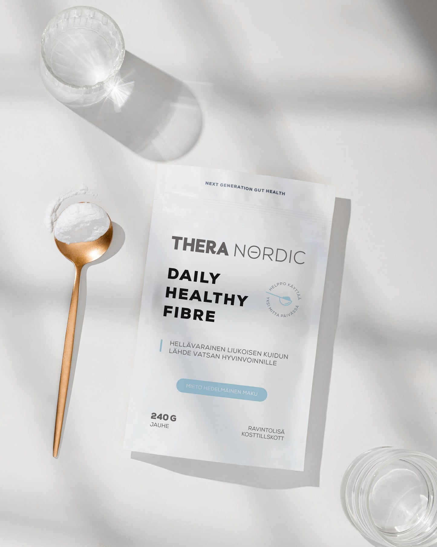 THERA Nordic Daily Healthy Fiber Kuitujauhe; valkoinen, mattapintainen pussi, jossa lukee "Thera Nordic Daily Healthy Fibre", asetettu sileälle, vaalealle pinnalle; etiketin teksti on groteskia, mustaa ja sinistä; vieressä kirkas vesilasi, pyöreä läpinäkyvä lasikulho ja vaalea puulusikka, jossa hienoa luonnonvalkoista jauhetta; kuvassa ei näy ihmisiä tai käsiä; pehmeitä varjoja minimalistisessa pöytäasetelmassa.