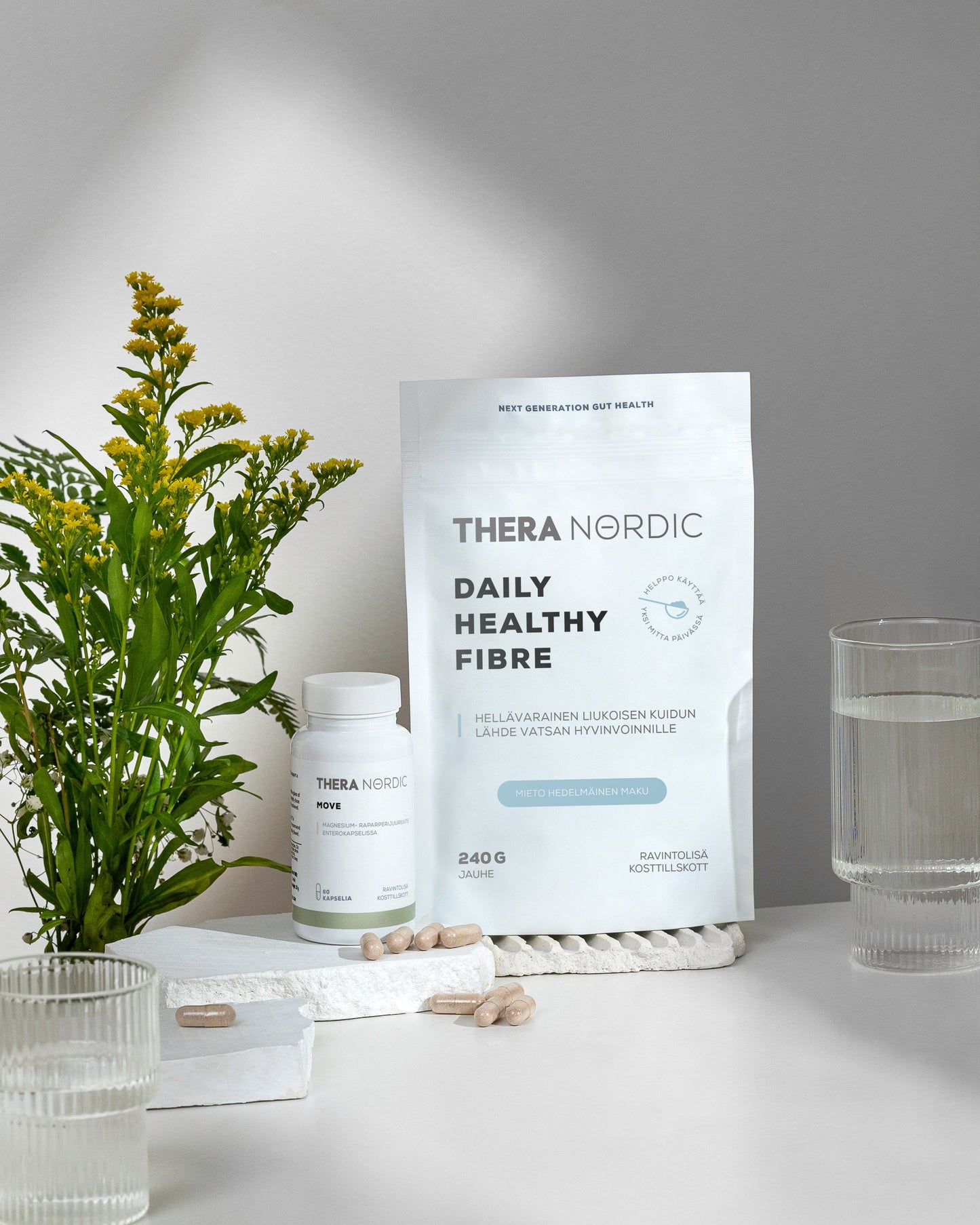 THERA Nordic Daily Healthy Fiber Kuitujauhe; pussi, jossa lukee "THERA NORDIC Daily Healthy Fibre", valkoisella korkilla varustettu pullo, beigejä kapseleita, kaksi vedellä täytettyä läpinäkyvää lasia, taitettu valkoinen pyyhe ja keltaisten kukkien kimppu on aseteltu sileälle valkoiselle pinnalle pehmeässä, hajautetussa valaistuksessa; kuvassa ei näy käsiä tai ihmisiä; kaikki esineet näkyvät kokonaan kuvassa.