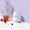 THERA Nordic Optimized Enzymes; sylinterinmuotoinen muovipullo valkoisella korkilla, jossa lukee lihavalla, modernilla groteskifontilla "THERA NORDIC Optimized Enzymes"; pullon vieressä läpinäkyvä lasi, jossa läpikuultamatonta vaaleanpunaista nestettä; sileällä, mattavalkoisella pinnalla vadelmia ja mustikoita; tausta tasaisen violetti; kuvassa ei näy ihmisiä tai käsiä.