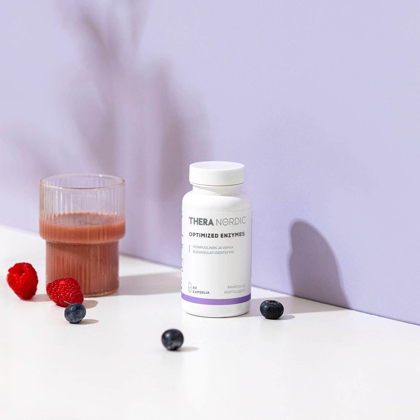 THERA Nordic Optimized Enzymes; sylinterinmuotoinen muovipullo valkoisella korkilla, jossa lukee lihavalla, modernilla groteskifontilla "THERA NORDIC Optimized Enzymes"; pullon vieressä läpinäkyvä lasi, jossa läpikuultamatonta vaaleanpunaista nestettä; sileällä, mattavalkoisella pinnalla vadelmia ja mustikoita; tausta tasaisen violetti; kuvassa ei näy ihmisiä tai käsiä.