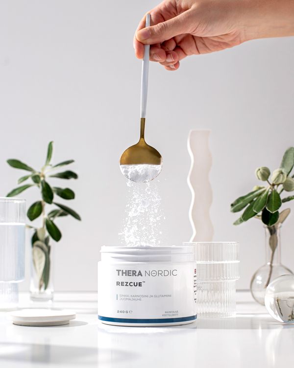 THERA Nordic Rezcue; käsi pitelee lusikkaa sylinterinmuotoisen, mattavalkoisen purkin yläpuolella, jossa lukee lihavalla groteskilla "THERA NORDIC"; jauhetta valuu lusikasta avoimeen purkkiin; kuva-ala on sileä valkoinen pinta, taustalla epätarkkoina viherkasveja ja kirkasta lasitavaraa; kuvassa ei näy muita ihmisiä kuin käsi.