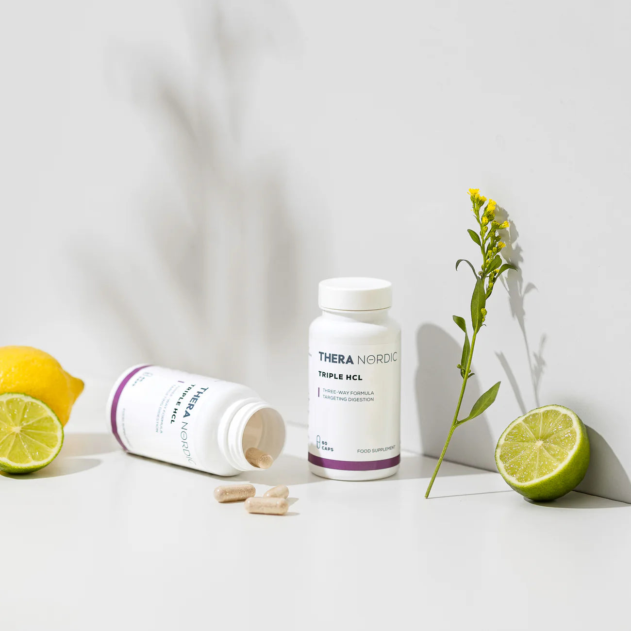 THERA Nordic Triple HCL Suolahappo; kaksi Thera Nordic -lisäravinnepulloa mattavalkoisella pinnalla ja sinisellä etiketillä, sylinterinmuotoiset ja valkoisin korkein; lähellä irtotabletteja; kuvassa myös kokonainen sitruuna, halkaistu lime ja keltainen kukka; etiketeissä siisti groteskitypografia ja luettava tuotenimi; tausta on tasainen valkoinen pinta pehmein varjoin luonnonvalossa.