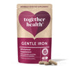 A matte pouch labeled Together Health Gentle Iron Wholefood Supplement with a heart design is shown upright beside three beige capsules. The packaging is light-colored, rectangular with a seal at the top, and features simple sans-serif typography. Only label text legible: product name and brief supplement information. Plain white background, no people or objects present.