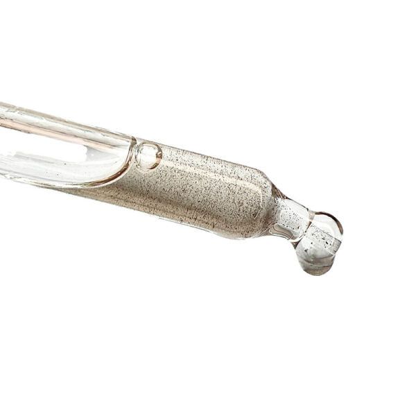 A close-up of a glass dropper with hyaluronic acid and tiny suspended particles, with a droplet forming at the tip, against a white background. The dropper is transparent and cylindrical, made of glass with a glossy finish. No label or text is visible on the product. The shot does not include hands or any other objects in the frame.