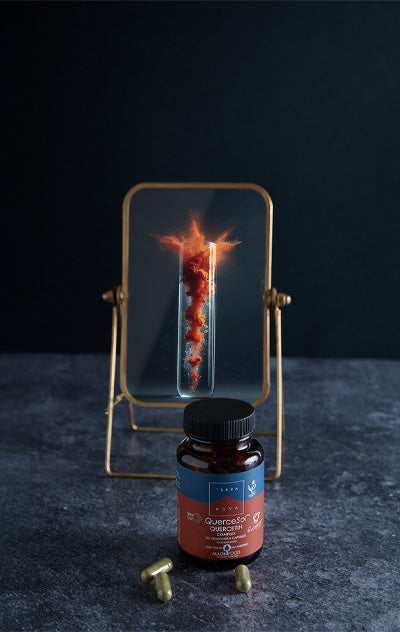Terranova NextGen Quercesol Quercetin Complex; ruskea muovinen ravintolisäpullo tekstillä ”Quercetin” tasaisella harmaalla mattapinnalla; useita vaaleansävyisiä kapseleita aseteltuna pullon juurelle; etiketissä yksinkertaista, selkeää typografiaa; taustalla läpinäkyvä lasikehys näyttää kuvan koeputkesta, josta nousee oranssinpunainen pilvi; ei ihmisiä tai käsiä kuvassa.