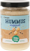 Terrasana Luomu Hummus; lasipurkki luomuhummusta mustalla kannella; etiketti on sininen ja beige mattapinnalla, kääritty sylinterinmuotoisen kirkkaan lasipurkin ympärille; etuosassa kuvituksia kikherneistä ja seesaminsiemenistä; groteskitekstiä tuotetiedoissa, näkyvissä maininta "positive eating"; tausta on yksivärinen valkoinen, ei ihmisiä tai muita esineitä näkyvissä.
