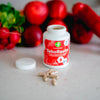 Terveyskaista TehoRauta 100 mg: valkoinen, mattapintainen sylinterimäinen muovipullo ruuvikannella seisoo avoimena tasaisella keittiötasolla; etiketissä teksti "TehoRauta" lihavoidulla tummalla groteskikirjasimella; pieniä ruskeita rautakapseleita on levinnyt pullon ympärille; taustalla epätarkkoja punaisia hedelmiä ja vihanneksia.
