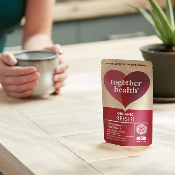 Together Health Reishi; mattapintainen beige pussi tekstillä Together Health Organic Reishi seisoo pystyssä puupöydällä; pieni selkeä groteskiteksti näkyvissä pakkauksessa; vieressä pieni vihreä lehtikasvi; epätarkassa taustassa henkilö pitelee mukia molemmin käsin rintakorkeudella; sisätila luonnollisessa valaistuksessa.