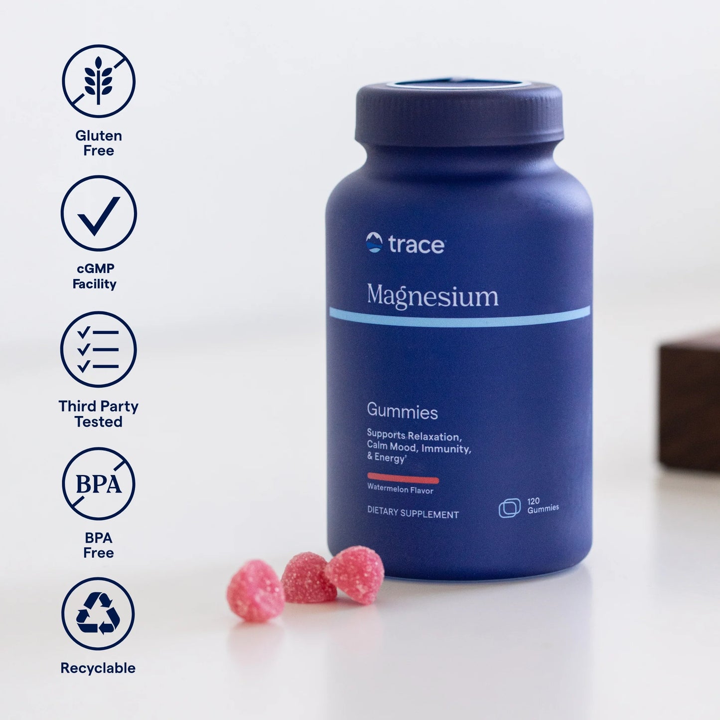 Trace Minerals Magnesium Gummies Watermelon; sininen sylinterinmuotoinen muovipullo, jossa lukee ”trace Magnesium Gummies”, mattapinnalla, on aseteltu valkoiselle tasaiselle pinnalle; lähellä kolme vaaleanpunaista, kuvunmuotoista purukumia; etiketissä selkeää groteskitypografiaa; pullon vieressä pyöreitä kuvakkeita gluteenittomuudesta, cGMP-laitoksesta, kolmannen osapuolen testauksesta, BPA-vapaudesta ja kierrätettävyydestä; ei ihmisiä tai käsiä kuvassa.