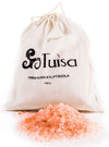 A cloth bag labeled Tuisa Himalayan Kylpysuola 1000g sits behind a pile of pure pink Himalayan bath salt crystals spread out in front. The bag is off-white cotton with a drawstring, features simple black printed text, and has a matte surface. The bath salt crystals are irregular chunks, pale to medium pink. Background is plain white; no people or props visible.