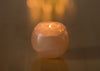 A lit candle burns inside a round, pink Himalayan salt holder, casting a soft, warm glow on a smooth brown surface. The holder is solid and has a matte finish. There is no visible label or packaging. The background and setting are minimal with only the surface visible, and there are no people or hands in the shot.