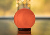 A glowing orange-red sphere sits on a reflective surface, with a blurred warm background and small bokeh lights visible behind it. The sphere appears glossy, with no visible label or markings. There is no packaging or text present. No people, hands, or actions are visible in the shot. The setting is indoors and the image focuses tightly on the product.