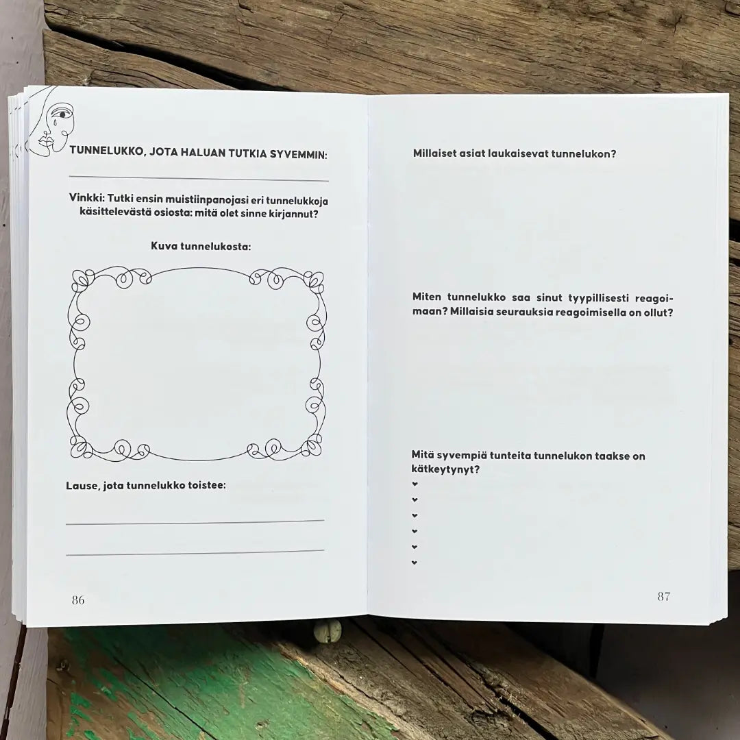 Tutustu tunnelukkoihin; työkirja avattuna sivuille, joilla suomenkielistä tekstiä, koristeellisia reunuksia ja kirjoitustehtäviä tunteista; kirja lepää rustiikkisella puupinnalla vihrein ja luonnollisen ruskein lautoin; mattapintaiset sivut valkoisia mustalla painetulla tekstillä ja käsinpiirretyn tyylisin reunuksin; sivunumerot 86 ja 87 näkyvissä alakulmissa; ei ihmisiä tai käsiä kuvassa.