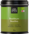A green tin of Urtekram organic basil, labeled in multiple languages, features a black lid and contains 20 grams of aromatic herb. The metal container is cylindrical with a matte finish and has simple printed text on the front in sans-serif fonts. The tin sits upright against a plain white background with no other objects or people visible in the image.