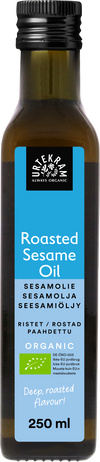 Urtekram Luomu Paahdettu Seesamiöljy, pullo luomupaahdetulle seesamiöljylle sinisellä etiketillä, tuotenimi näkyy usealla kielellä ja tilavuus 250 ml, korostaen premium-laatua; pullo sylinterimäinen, tummaa lasia mattapinnalla ja kierrekorkilla; sinisessä etiketissä valkoista groteskitekstiä ja vähäisiä graafisia elementtejä; tausta valkoinen, ei ihmisiä eikä käsiä näkyvissä; suoraan edestä otettu tuotekuva.