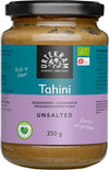Urtekram Luomu Suolaton Tahini, sylinterimäinen lasipurkki luomu-suolattomalle tahinille violetilla mattaetiketillä, mustalla muovisella kierrekannella ja kirkkaalla rungolla, jonka läpi näkyy vaaleanbeigeä sisältöä; etiketissä lihavoitua valkoista ja vihreää tekstiä: "Rich in fiber!" ja "Source of protein!" sekä tuotetietoja ja ravintosisältötietoja usealla kielellä; ei ihmisiä eikä toimintaa näkyvissä; kuva otettu yksinkertaista taustaa vasten.