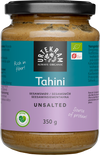 Urtekram Luomu Suolaton Tahini, sylinterimäinen lasipurkki luomu-suolattomalle tahinille violetilla mattaetiketillä, mustalla muovisella kierrekannella ja kirkkaalla rungolla, jonka läpi näkyy vaaleanbeigeä sisältöä; etiketissä lihavoitua valkoista ja vihreää tekstiä: "Rich in fiber!" ja "Source of protein!" sekä tuotetietoja ja ravintosisältötietoja usealla kielellä; ei ihmisiä eikä toimintaa näkyvissä; kuva otettu yksinkertaista taustaa vasten.