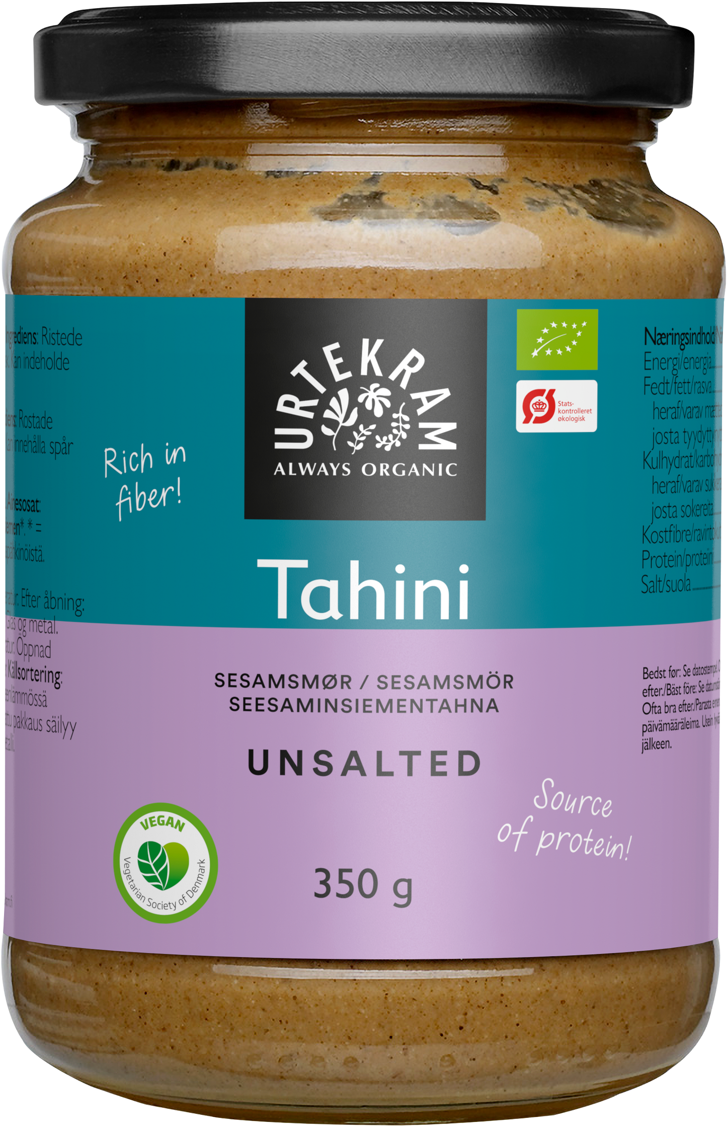 Urtekram Luomu Suolaton Tahini, sylinterimäinen lasipurkki luomu-suolattomalle tahinille violetilla mattaetiketillä, mustalla muovisella kierrekannella ja kirkkaalla rungolla, jonka läpi näkyy vaaleanbeigeä sisältöä; etiketissä lihavoitua valkoista ja vihreää tekstiä: "Rich in fiber!" ja "Source of protein!" sekä tuotetietoja ja ravintosisältötietoja usealla kielellä; ei ihmisiä eikä toimintaa näkyvissä; kuva otettu yksinkertaista taustaa vasten.