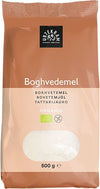 A 500g bag of Urtekram organic buckwheat flour with multilingual labeling and a transparent window displaying the flour inside. The packaging is upright, rectangular, and made of matte paper with green accents. The label features bold sans-serif text, including weight and product name. Photo background is plain and light-colored. No people or hands are visible.