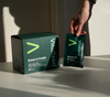 Vitalized Greens & Protein; käsi pitelee pussia tekstillä Vitalized Greens & Protein vastaavan tuotelaatikon vieressä valkoisella pinnalla, auringonvalo luo varjoja ja korostaa tuoreita ainesosia; pussi ja laatikko suorakaiteen muotoisia mattapinnalla vihreä-valkoisin värein; molemmissa selkeää groteskitypografiaa näkyvässä etikettitekstissä; vain yksi käsi näkyvissä, ei muita ihmisiä tai esineitä paitsi lähellä aseteltuja tuoreita vihreitä kasveja; lähikuva luonnonvalossa.