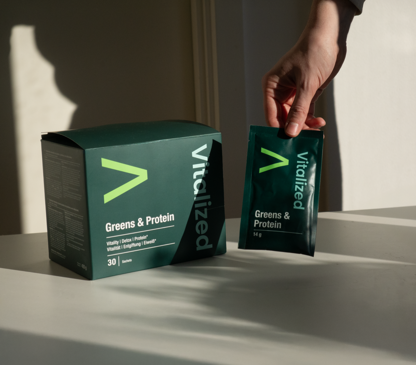 Vitalized Greens & Protein; käsi pitelee pussia tekstillä Vitalized Greens & Protein vastaavan tuotelaatikon vieressä valkoisella pinnalla, auringonvalo luo varjoja ja korostaa tuoreita ainesosia; pussi ja laatikko suorakaiteen muotoisia mattapinnalla vihreä-valkoisin värein; molemmissa selkeää groteskitypografiaa näkyvässä etikettitekstissä; vain yksi käsi näkyvissä, ei muita ihmisiä tai esineitä paitsi lähellä aseteltuja tuoreita vihreitä kasveja; lähikuva luonnonvalossa.