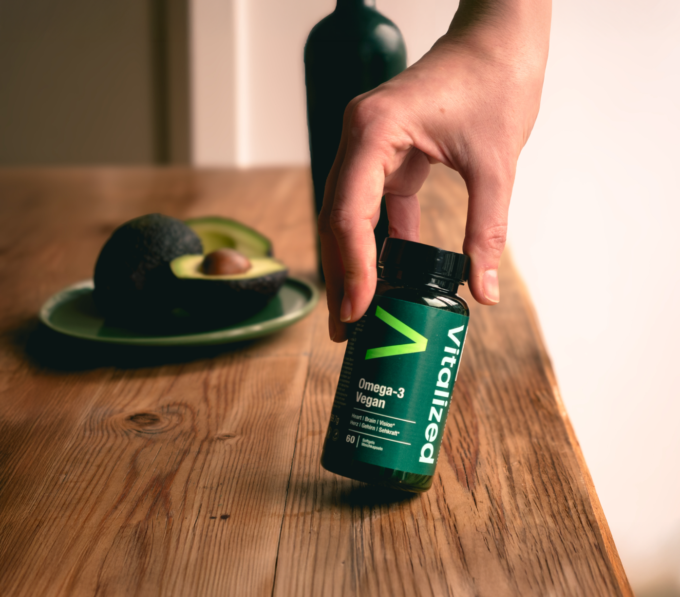 Vitalized Omega-3 Vegan; käsi pitelee valkoista muovipulloa tekstillä Vitalizea Omega-3 Vegan groteskifontilla mattapintaisella puupöydällä; taustalla pyöreä lautanen tasaisesti viipaloitua avokadoa ja tumma lasipullo kiiltäväpinnalla; kohtaus hyvin valaistu, kuvattu ylhäältä.