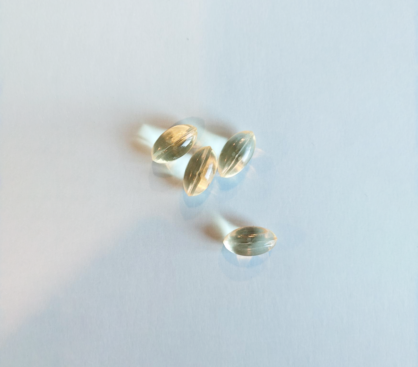 Vitalized Omega-3 Vegan; neljä kirkasta, soikeanmuotoista geelikapselia aseteltuna tasaiselle valkoiselle pinnalle, niiden kiiltävä pinta luo pehmeitä varjoja; kapselit läpinäkyviä ilman näkyvää sisältöä tai etikettejä; ei pakkausta kuvassa; asetelma minimalistinen ilman taustaelementtejä, tekstiä, ihmisiä, käsiä tai muita esineitä.