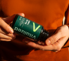 Vitalized SAMe+ 200 mg; henkilö oranssissa paidassa pitelee vihreää muovipulloa kiiltäväpinnalla; etiketti näytetty kulmassa luettavuuden vuoksi, esittää sanat Vitralized, 200mg ja Shine+ selkeällä modernilla typografialla; ei muita pakkaus- tai suunnitteluelementtejä näkyvissä; neutraali tausta, tuote kädessä, keskikokoinen lähikuva.