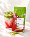 Lasi vaaleanvihreää ja vaaleanpunaista matcha-mansikkalattea on Vivani Matcha Strawberry -suklaapatukan vieressä, jonka mattapintaisessa kääreessä on vihreitä ja kirkkaan pinkkejä osioita. Vivani-logo ja tuotenimi näkyvät mustalla pääteviivattomalla fontilla. Tuotteet on aseteltu puhtaalle valkoiselle alustalle, etualalla on ripoteltuna elävää vihreää matchajauhetta. Ihmisiä tai käsiä ei näy.