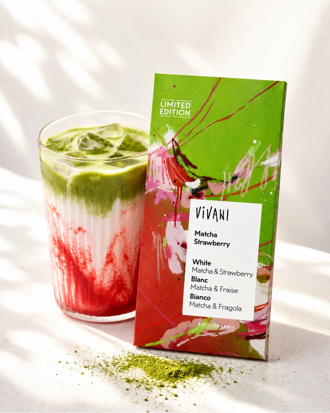 Lasi vaaleanvihreää ja vaaleanpunaista matcha-mansikkalattea on Vivani Matcha Strawberry -suklaapatukan vieressä, jonka mattapintaisessa kääreessä on vihreitä ja kirkkaan pinkkejä osioita. Vivani-logo ja tuotenimi näkyvät mustalla pääteviivattomalla fontilla. Tuotteet on aseteltu puhtaalle valkoiselle alustalle, etualalla on ripoteltuna elävää vihreää matchajauhetta. Ihmisiä tai käsiä ei näy.