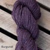 A single, twisted skein of deep purple yarn rests on light-colored wooden planks. The yarn has a matte finish and is unwrapped, showing the fiber texture. The photo is taken from overhead. “Burgundi” appears in simple, lowercase black type in the bottom-left corner. No people or packaging are visible.