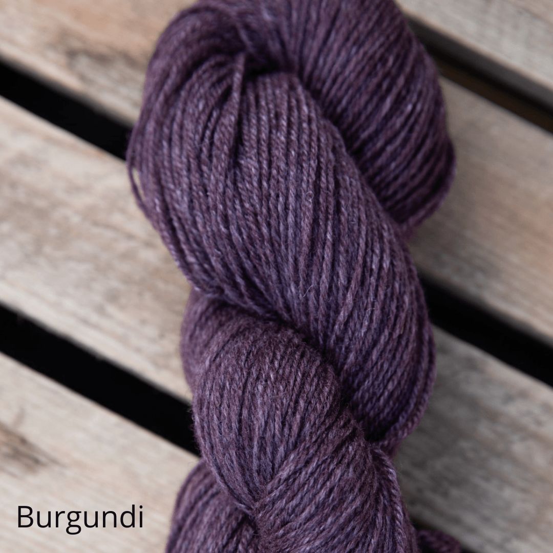 A single, twisted skein of deep purple yarn rests on light-colored wooden planks. The yarn has a matte finish and is unwrapped, showing the fiber texture. The photo is taken from overhead. “Burgundi” appears in simple, lowercase black type in the bottom-left corner. No people or packaging are visible.