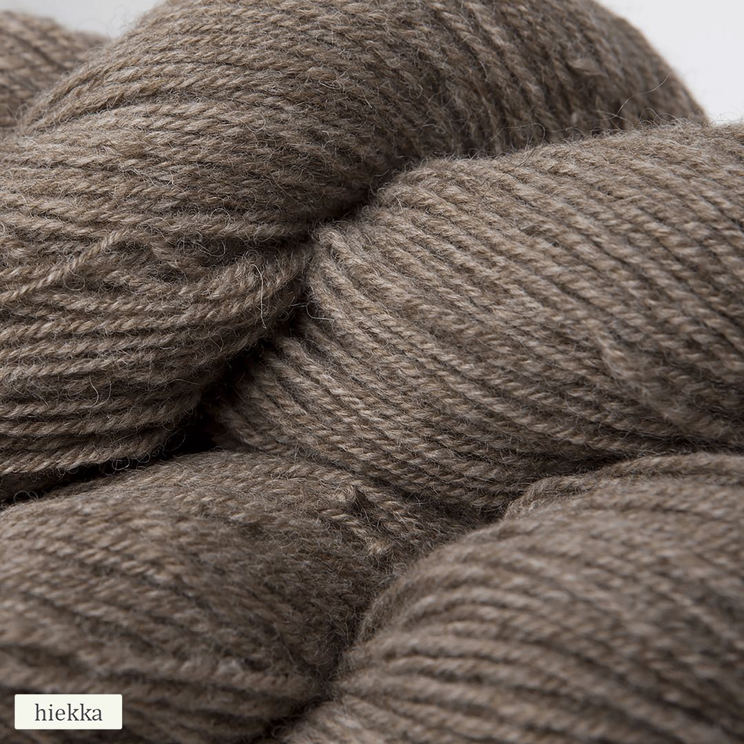 Close-up of three skeins of brownish-gray alpaca yarn with a matte, soft texture. Each skein is loosely wound and cylindrical. A small label in the lower left corner says hiekka in simple, sans-serif font. The background is plain and neutral. No packaging visible other than the label. No people or hands shown.