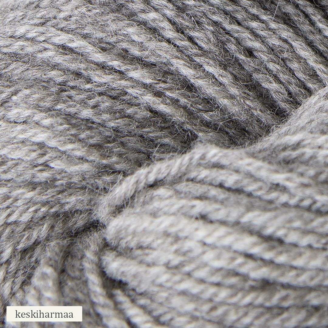Close-up of gray yarn with a soft texture, showing its twisted fibers. The yarn is matte, loosely wound, and fills most of the frame. A small white label in the bottom left corner displays the word “keskiharmaa” in simple, black sans-serif text. There is no packaging visible. The background appears neutral and uncluttered.