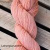 A close-up of a skein of salmon pink yarn, also known as Lohenpunainen, placed on a wooden surface. The yarn is matte and wound into an oval shape. There is no visible packaging. The background is light-colored wood with distinct grain patterns. The word Lohenpunainen appears in the lower left corner in simple, sans-serif text. No people or hands are present.