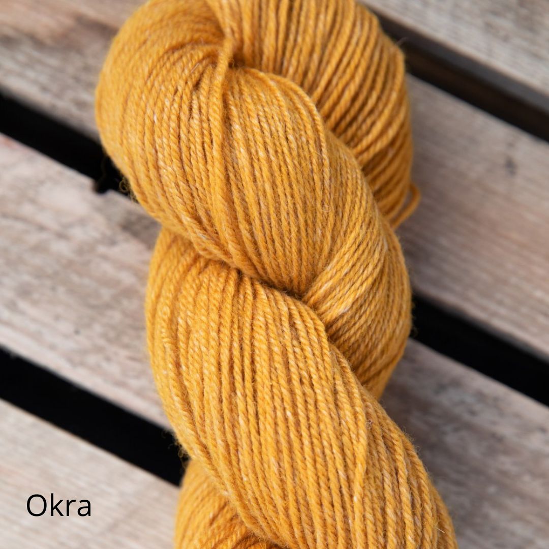 A skein of golden-yellow yarn labeled Okra, spun from soft fibers, rests on a wooden surface. The yarn is wound into an oblong skein with a matte finish. The label features the word “Okra” in clear, sans-serif type. No packaging beyond the label is present. The background is a plain, medium-toned wood grain seen from above. No people or hands are visible in the shot.