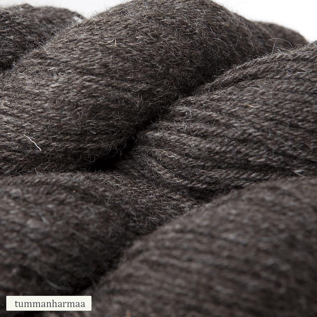 Close-up of dark gray wool yarn with a slightly coarse texture. The material is matte with visible fibers and a round, bundled form. A small white label in the corner shows plain, sans-serif text: tummanharmaa. Background is neutral and uncluttered. No hands or people visible in the image.