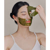 Whamisa Algae Kelp Mask; valkoisessa hihattomassa paidassa oleva henkilö kuorii hellästi kiiltävää, vihreää kangasnaamiota kasvoiltaan, silmät kiinni; tausta vaalea ja tasainen ilman näkyvää pakkausta tai brändäystä; ei etikettitekstiä tai typografiaa kuvassa; kuva keskittyy henkilön yläkehoon ja hartioihin.