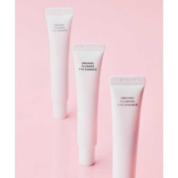 Whamisa Organic Flowers Eye Essence; kolme valkoista muovitupua mattapinnalla, tekstillä ”ORGANIC FLOWERS EYE ESSENCE” versaaligroteskilla; tuubit identtisiä ja pystyasennossa yksivärisellä vaaleanpunaisella alustalla; ei ihmisiä tai käsiä; sommittelu minimalistinen, pehmeäfokuksinen kuvaustyyli korostaa tuotteita tasaista taustaa vasten.