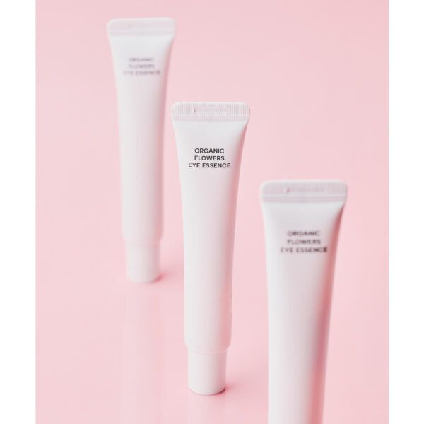 Whamisa Organic Flowers Eye Essence; kolme valkoista muovitupua mattapinnalla, tekstillä ”ORGANIC FLOWERS EYE ESSENCE” versaaligroteskilla; tuubit identtisiä ja pystyasennossa yksivärisellä vaaleanpunaisella alustalla; ei ihmisiä tai käsiä; sommittelu minimalistinen, pehmeäfokuksinen kuvaustyyli korostaa tuotteita tasaista taustaa vasten.