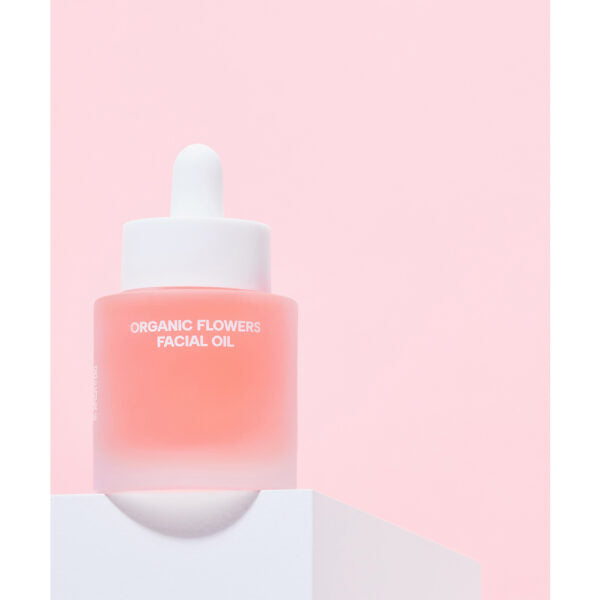 Whamisa Organic Flowers Facial Oil Deep Rich; huurrelasinen tippapullo tekstillä ”ORGANIC FLOWERS FACIAL OIL” lepää valkoisella alustalla pastellinvaaleanpunaista taustaa vasten; pullo lieriömäinen pyöristetyin olkaosin ja valkoinen kierrekorkki tipoittimineen; etikettiteksti versaaligroteskilla; ei ihmisiä tai käsiä; kuva pehmeässä, hajautetussa valaistuksessa yksinkertaisessa studioasetelmassa.