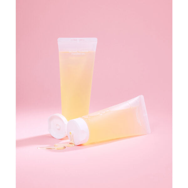 Whamisa Organic Flowers Foaming Gel; kaksi läpikuultavaa keltaista geelituubia valkoisin korkein, toinen pystyssä ja toinen vaakatasossa geelin valuessa suuttimesta, pastellinvaaleanpunaisella taustalla; tuubit lieriömäisiä ja kiiltäväpintaisia, ei näkyviä etikettejä tai tekstiä; ei ihmisiä tai muita esineitä kuvassa; keskikokoinen lähikuva tuotteesta.