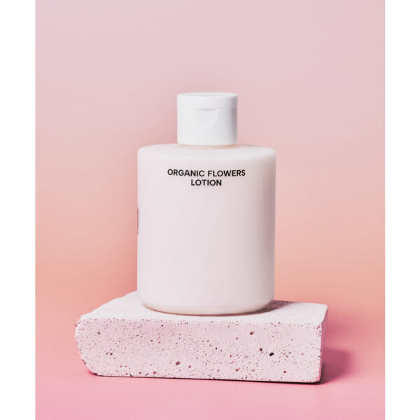 Whamisa Organic Flowers Lotion Double Rich; valkoinen pullo tekstillä ”ORGANIC FLOWERS LOTION” aseteltuna valkoiselle tekstuuripintaiselle korokkeelle pehmeää vaaleanpunaista liukuväritaustaa vasten, korostaen luonnonmukaista kauneutta; pullo lieriömäinen mattapinnalla ja yksinkertaisella mustalla groteskitekstillä; pakkaus minimalistinen ilman lisäkuvioita; kuva keskittyy tuotteeseen, ei ihmisiä tai käsiä.