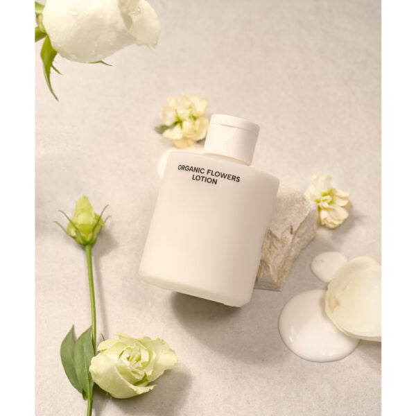 Whamisa Organic Flowers Lotion Double Rich; valkoinen muovipullo mattapinnalla, lieriömäinen muoto, painettu etiketti tekstillä ”ORGANIC FLOWERS LOTION” selkeällä groteskifontilla; pullo pystyasennossa sileällä vaalealla alustalla; ympärillä pieniä valkoisia kukkia ja epäsäännöllinen voideläikkä lähellä; ei ihmisiä tai käsiä kuvassa.