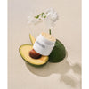 Whamisa Organic Flowers Nourishing Cream; kasvovoidepurkki aseteltuna halkaistun avokadon päälle valkoisen kukan kera beigellä neutraalilla alustalla, korostaen luonnollisia ainesosia; purkki pyöreä, lasia tai muovia mattapinnalla tai puolimatalla, kansi yksivärinen; ei ihmisiä tai käsiä; ei näkyvää etikettitekstiä; kuva asetelmatyylinen pehmeässä, tasaisessa valaistuksessa.
