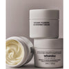 Whamisa Organic Flowers Nourishing Cream; kolme valkoista purkkia Whamisa Organic Flowers Nourishing Cream -voidetta; kaksi suljettua purkkia pinottuna, yksi avoinna näyttäen kermaisen, vaaleankeltaisen sisällön; purkit pyöreähköjä mattapinnalla; etiketeissä tuotenimi ja brändi mustalla tai harmaalla groteskitekstillä; tausta tasainen ja neutraali, ei ihmisiä tai käsiä; laaja tuotekuva.