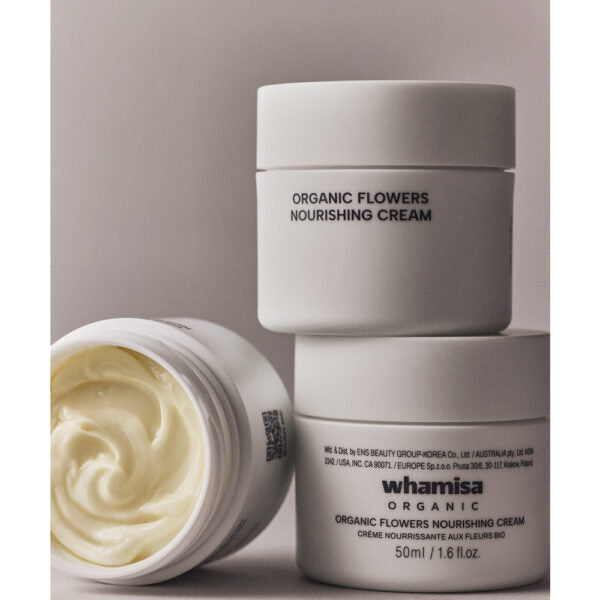 Whamisa Organic Flowers Nourishing Cream; kolme valkoista purkkia Whamisa Organic Flowers Nourishing Cream -voidetta; kaksi suljettua purkkia pinottuna, yksi avoinna näyttäen kermaisen, vaaleankeltaisen sisällön; purkit pyöreähköjä mattapinnalla; etiketeissä tuotenimi ja brändi mustalla tai harmaalla groteskitekstillä; tausta tasainen ja neutraali, ei ihmisiä tai käsiä; laaja tuotekuva.