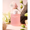 Whamisa Organic Flowers Toner Deep Rich; läpikuultavasta materiaalista valmistettu vaaleanpunainen pullo mattapinnalla, tekstillä ”Organic Flowers Toner” yksinkertaisella valkoisella groteskilla; pakkaus lieriömäinen pyöristetyllä olkaosalla ja valkoisella korkilla; aseteltuna valkoisten ja vihreiden kukkien sekä pienen puuosion joukkoon; tausta yksivärinen, pehmeän vaaleanpunainen; ei ihmisiä; kuva asetelmatyylinen edestä kuvattuna.